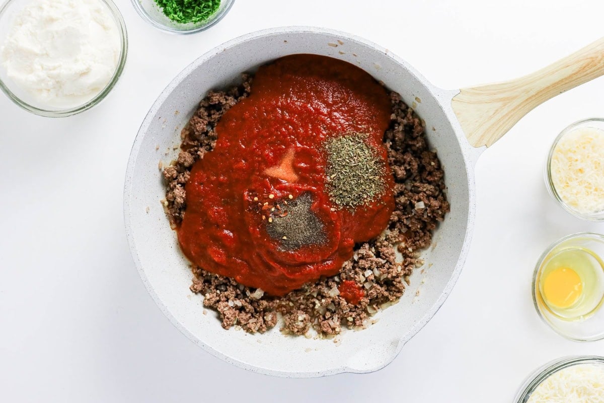 Skillet with browned ground beef topped with tomato sauce and herbs.