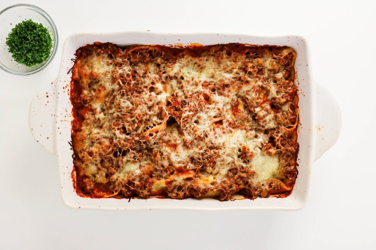 Baked pasta shells in a casserole dish next to a bowl of fresh herbs.