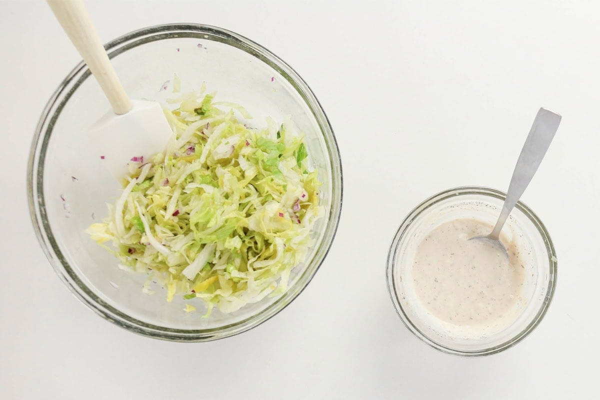Shredded lettuce in a bowl beside a bowl of creamy dressing on a white surface.