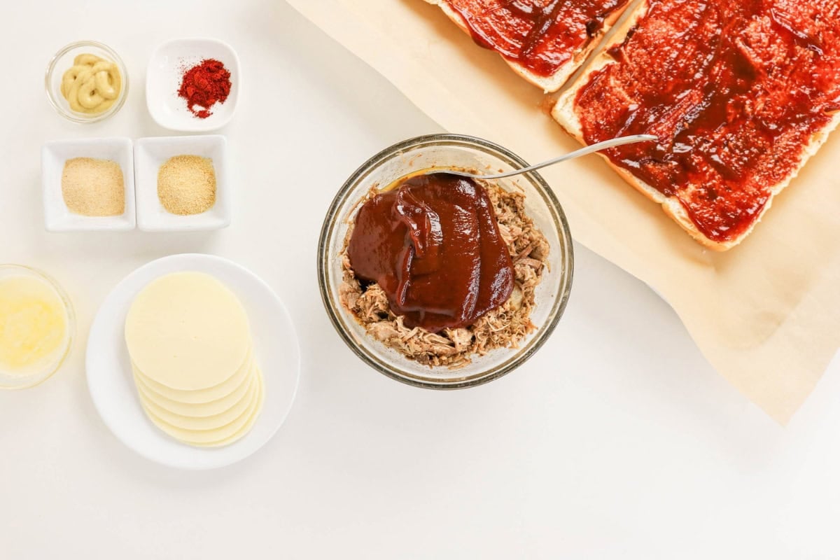 Bowl of BBQ pulled pork, cheese slices, spices, and sauced bread on a counter.