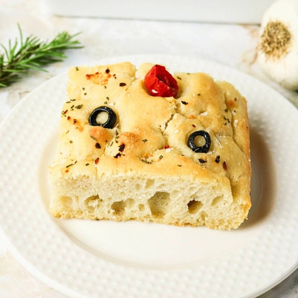 A slice of focaccia bread with olive, tomato, and herbs on a white plate.