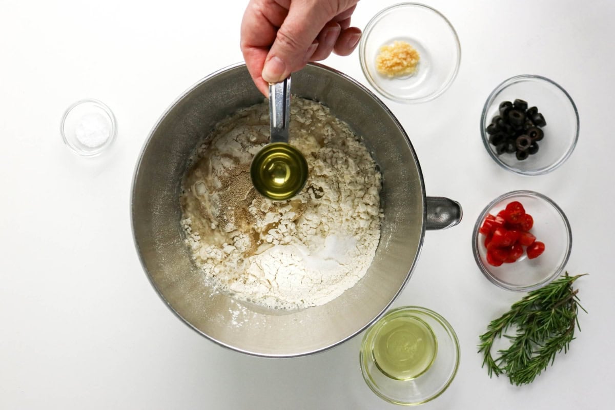 Oil is added to flour; nearby are olives, tomatoes, rosemary, and garlic.
