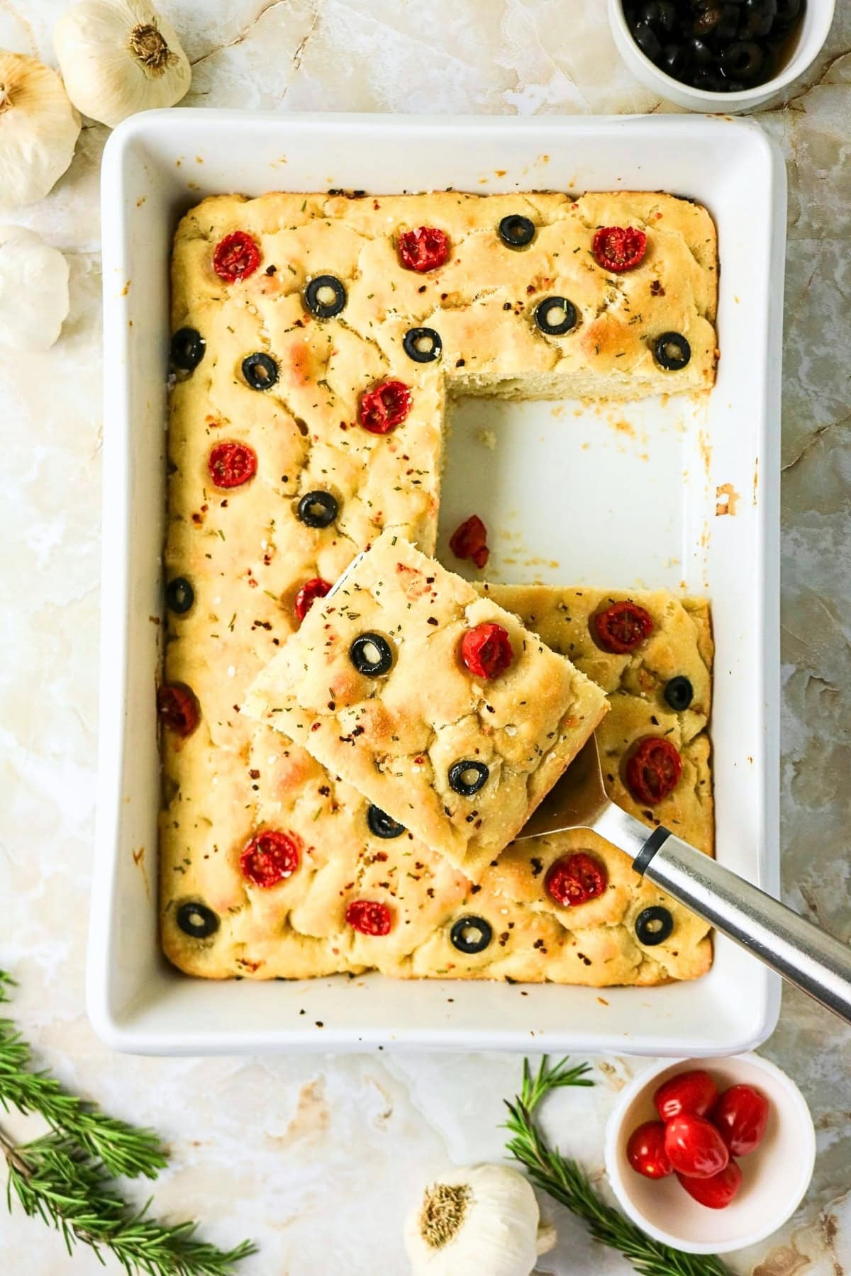 Focaccia in a pan with tomatoes and olives, one square being served.