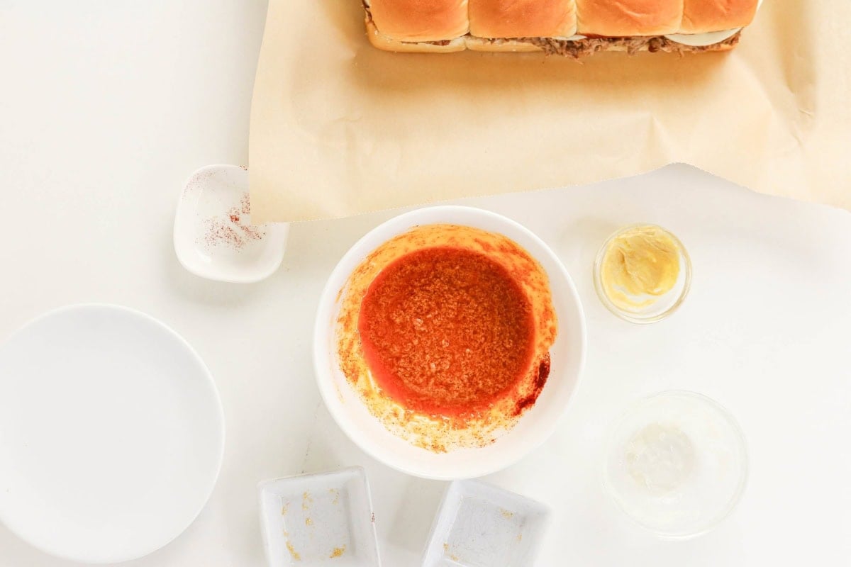 Bowl of orange sauce, mustard, sliders, and empty dishes on white surface.