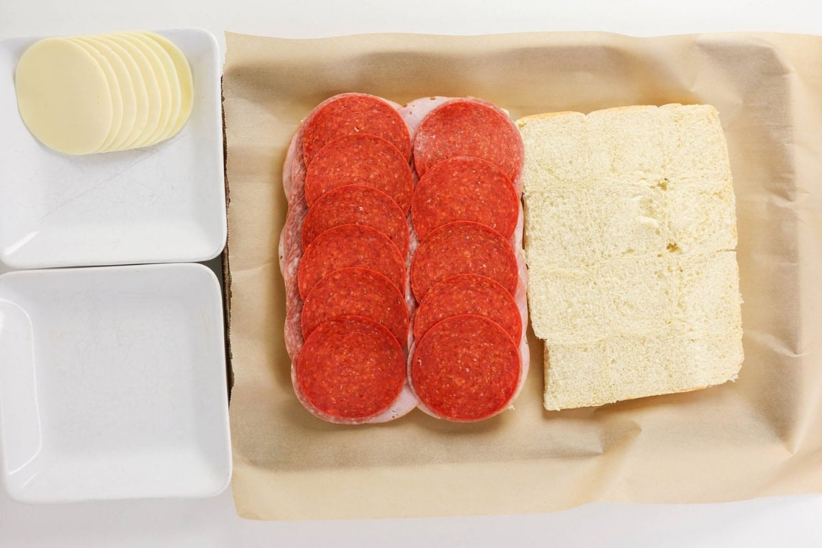Sliced cheese, pepperoni on meat, and bread on parchment and plates.