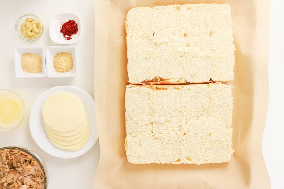 Sliced bread loaf with cheese, seasonings, and tuna on a white surface.
