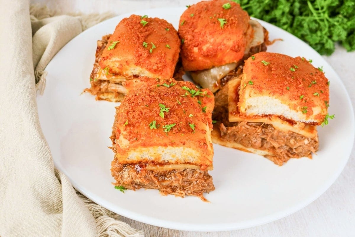 Four pulled pork sliders topped with sauce and parsley on a white plate.