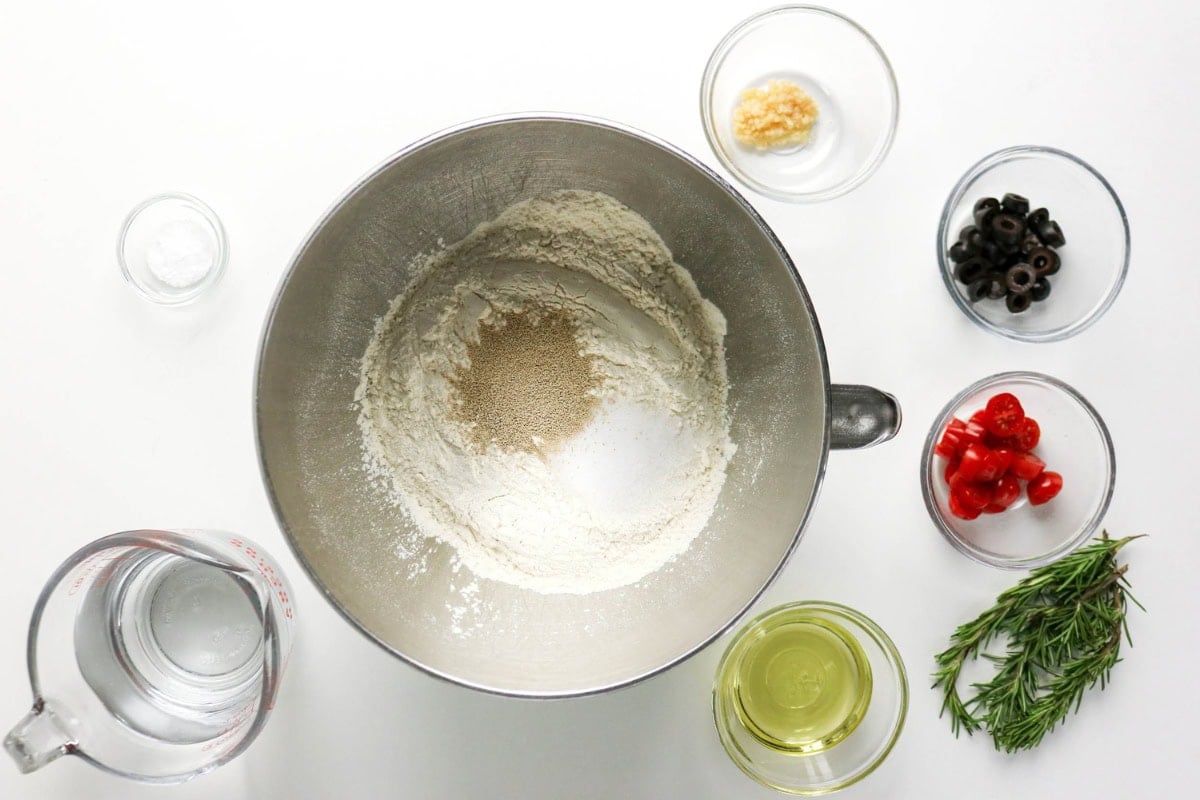 Mixing bowl with flour and yeast, surrounded by veggies, oil, water, and herbs.