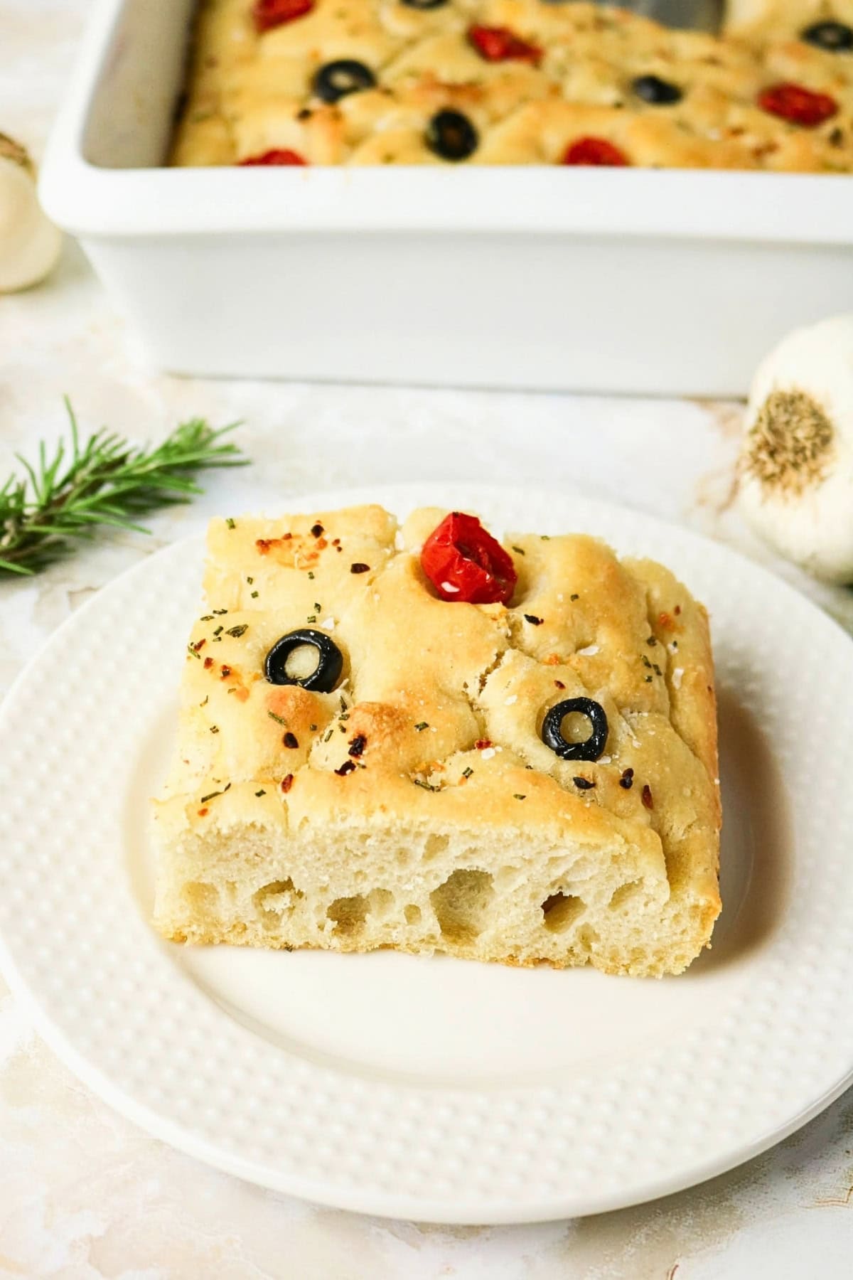 A slice of focaccia bread with olives and tomato on a white plate.