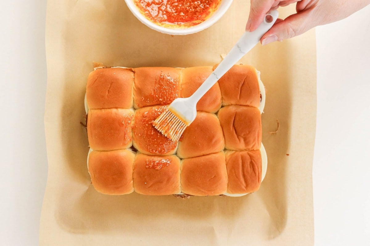 A hand brushes sauce on top of slider buns on parchment paper.