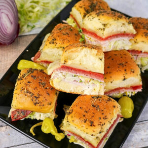 Mini sandwiches with lettuce, deli meats, and herbs on a black platter.