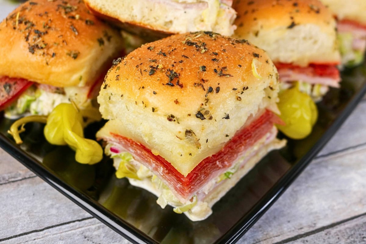 Mini sandwiches with deli meats and lettuce on a black plate, peppers on side.