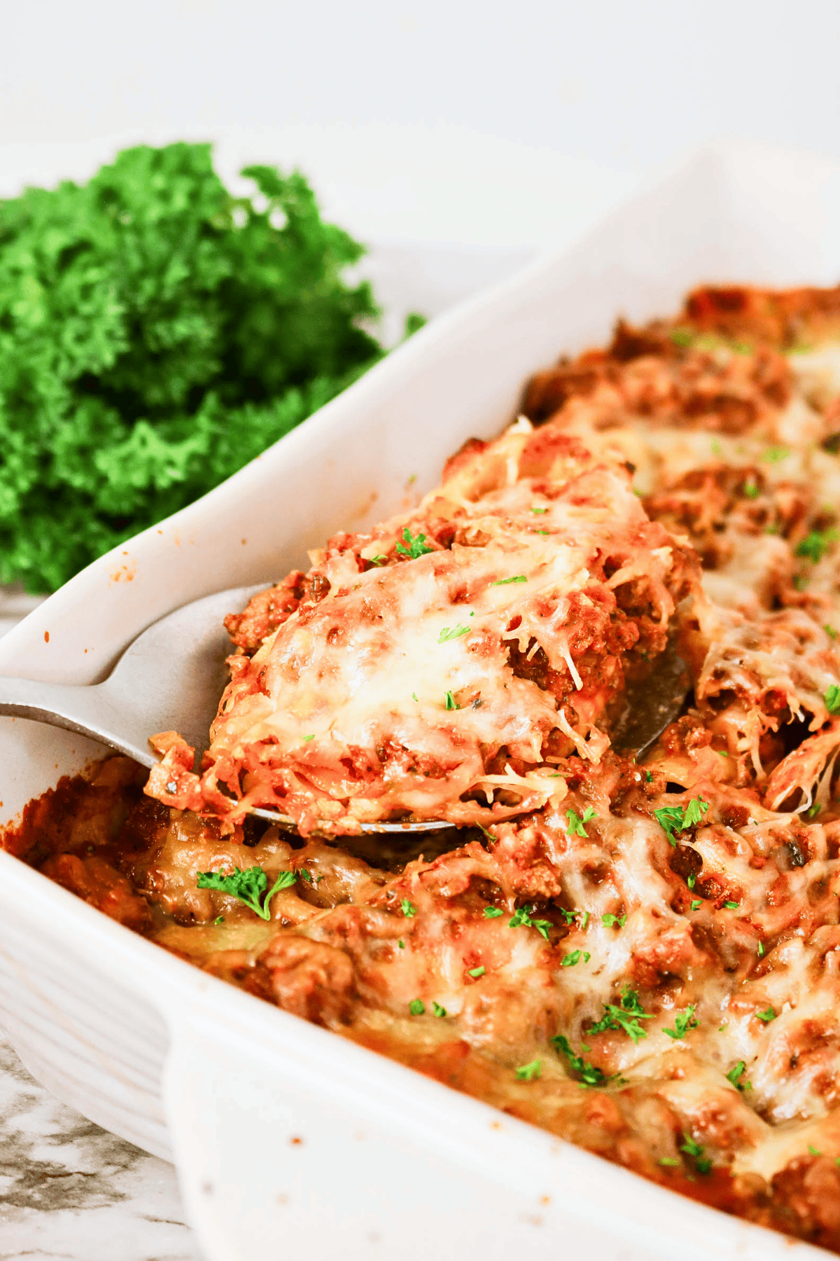 A serving spoon lifts a portion of cheesy baked pasta from a casserole dish.