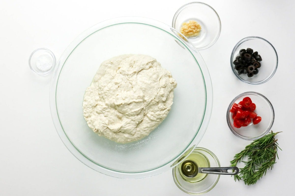 Dough in a glass bowl, with olives, tomatoes, rosemary, garlic, and oil nearby.