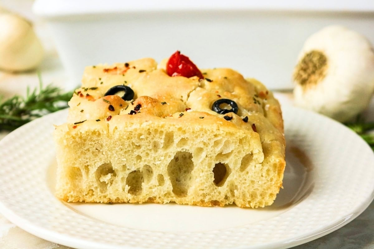 A slice of focaccia bread with olives and herbs on a white plate.