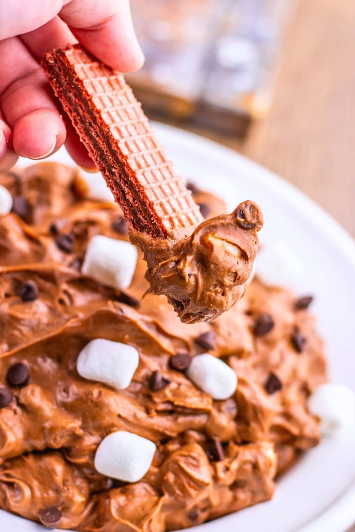 A hand dips a chocolate wafer into a bowl of chocolate dip with marshmallows.