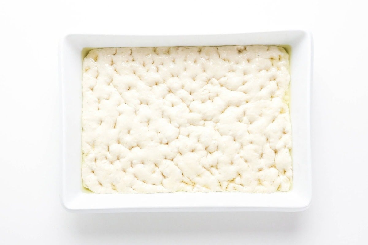 White baking dish with raw focaccia dough, dimpled and ready to bake.
