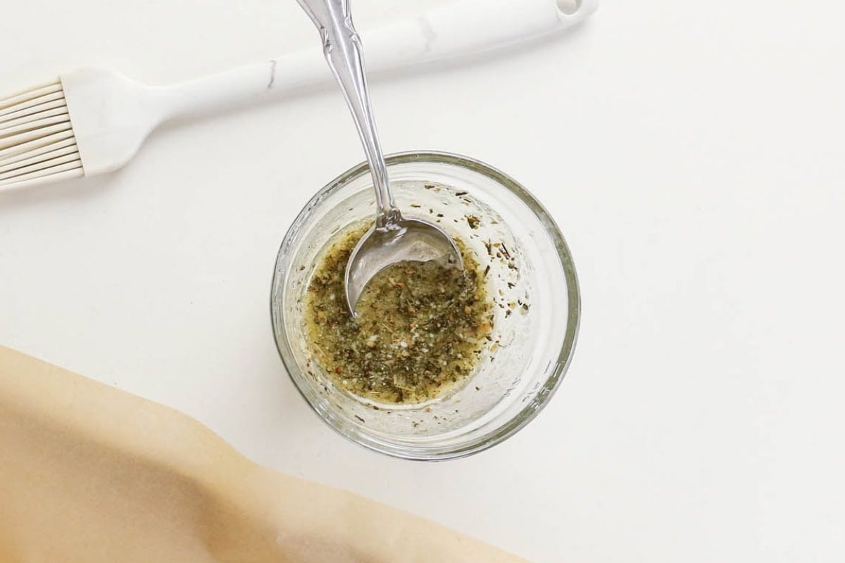 Small glass bowl with herb mixture and spoon, next to brush on white surface.
