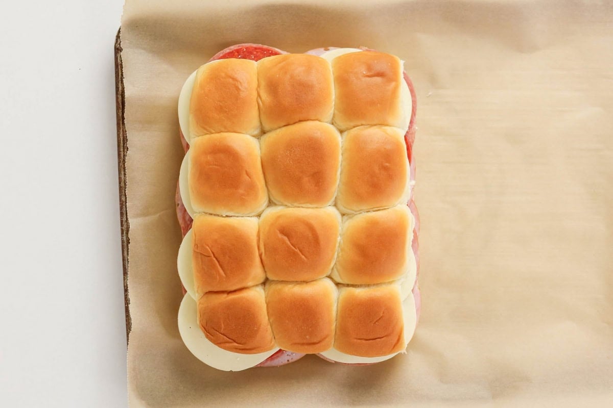 Twelve slider buns with cheese and meat on parchment paper.