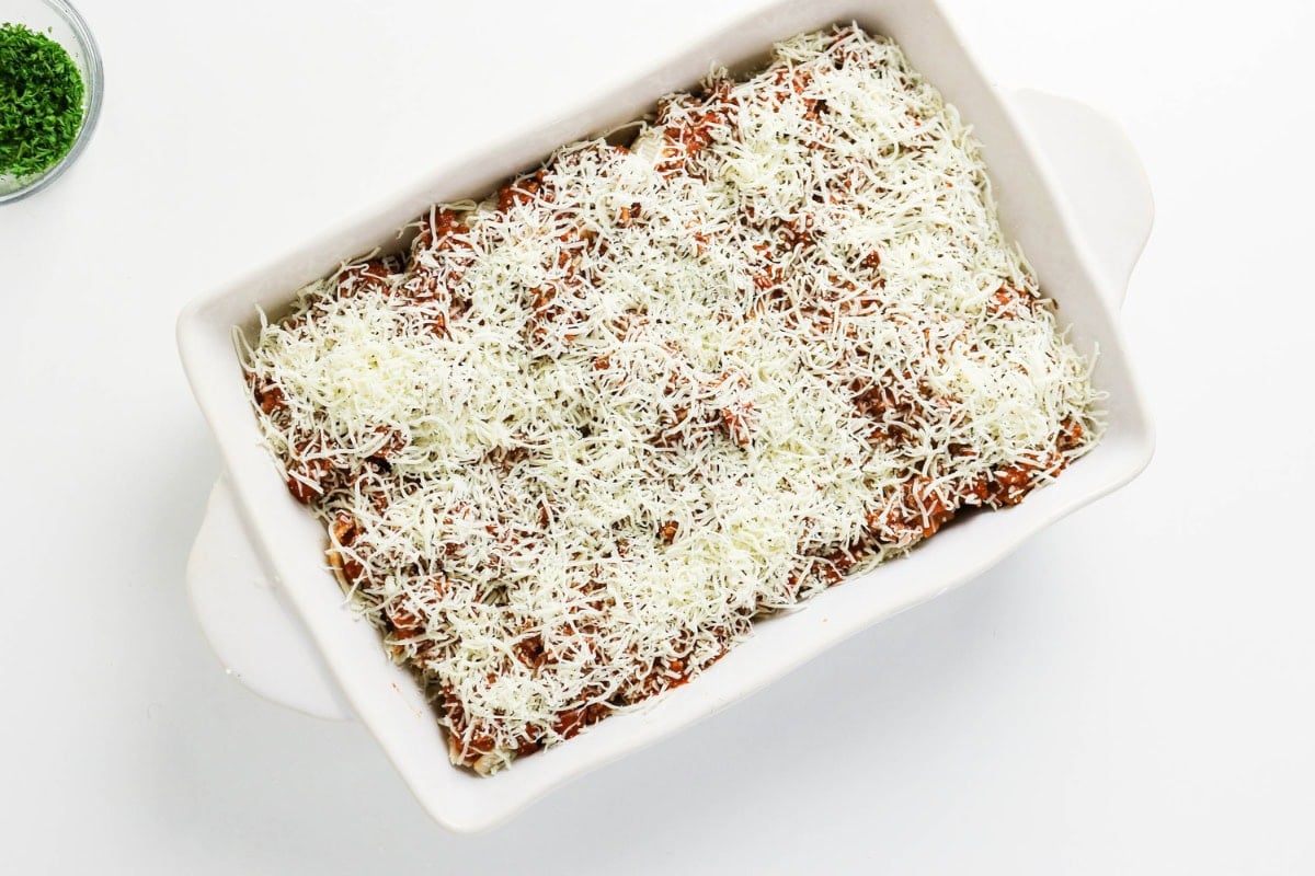 Unbaked lasagna with a layer of shredded cheese in a white baking dish.