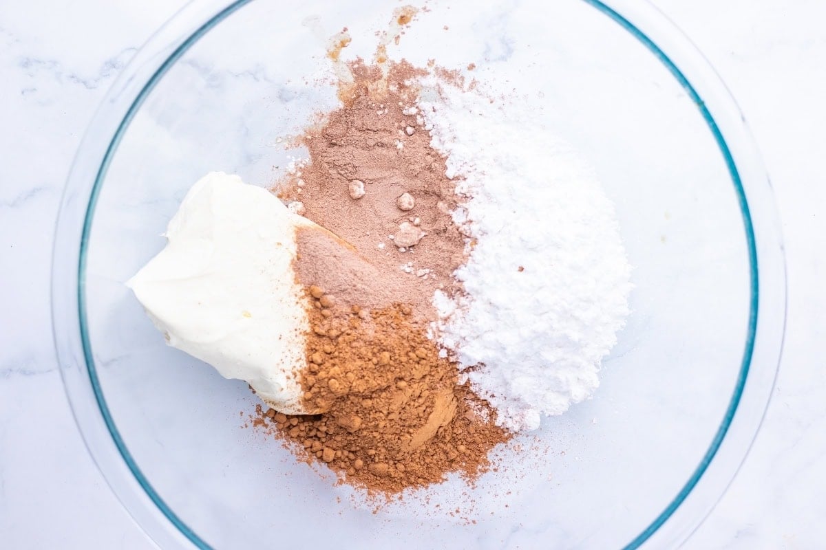 A glass bowl containing cream cheese, cocoa powder, and powdered sugar.