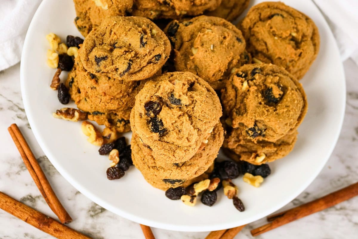 A stack of oatmeal raisin walnut cookies on a white plate with cinnamon sticks.