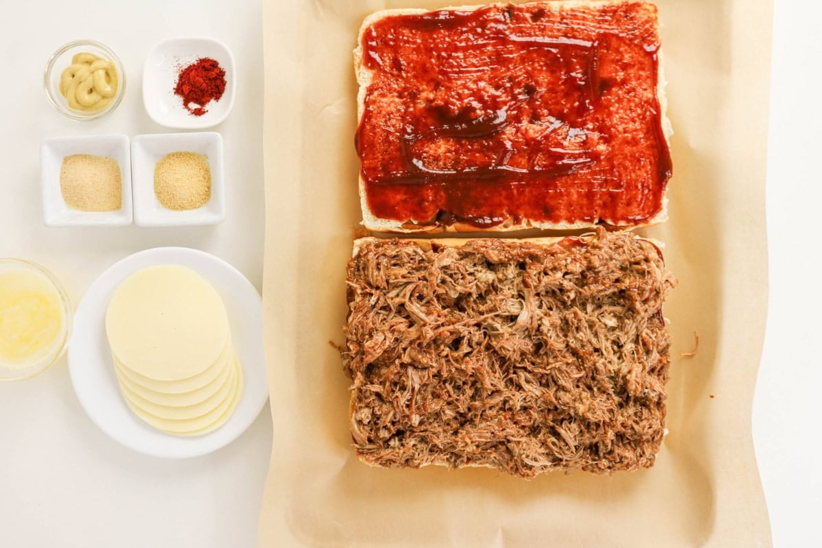 Two sandwich halves: one with BBQ sauce, one with shredded meat; sides nearby.