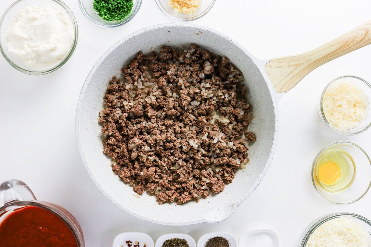 Ground beef and onions browning in a skillet surrounded by bowls of ingredients.
