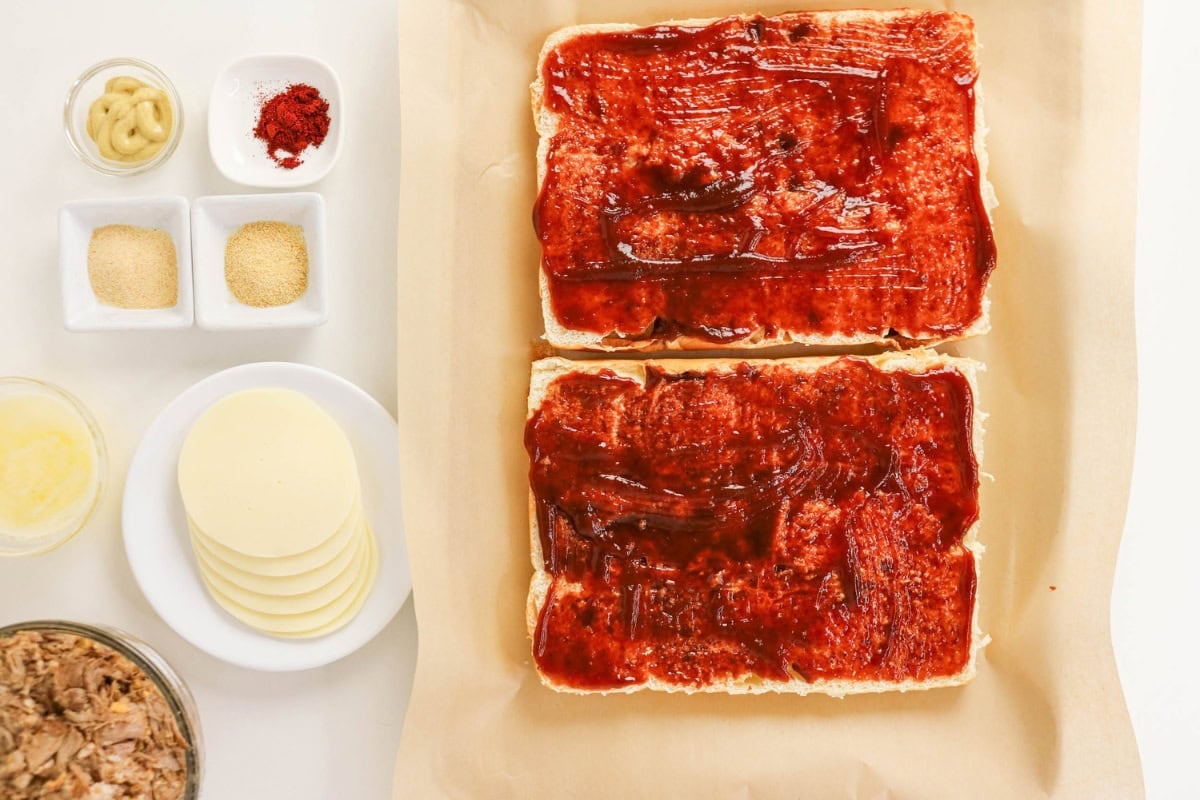 Two sandwich halves with sauce, cheese, seasonings, and tuna laid out nearby.