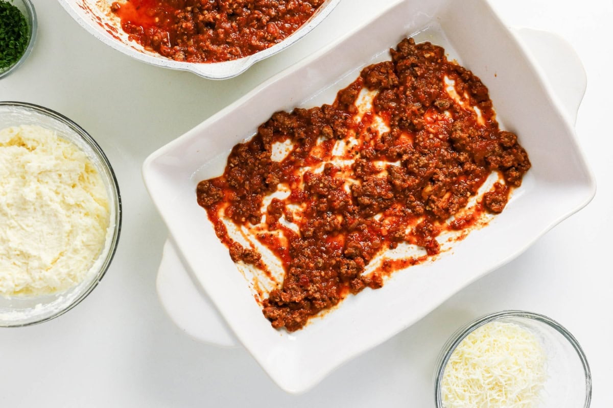A baking dish with a thin layer of meat sauce, next to bowls of cheese.